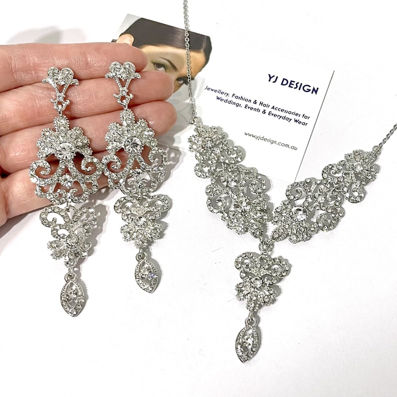 May include: A silver necklace and earrings set with a delicate, ornate design featuring clear rhinestones. The necklace has a pendant with a teardrop-shaped crystal.