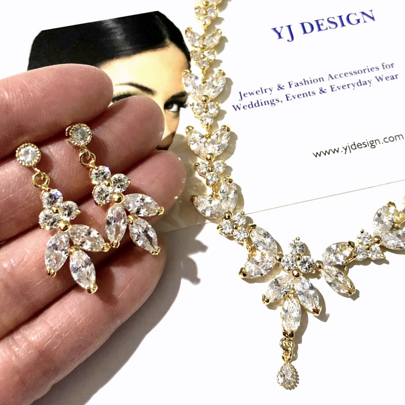 May include: Gold-toned jewelry set featuring a necklace and earrings. The necklace and earrings are adorned with clear, sparkling crystals and leaf-shaped accents. The earrings are dangling, and the necklace has a pendant. The text "YJ DESIGN" is visible.
