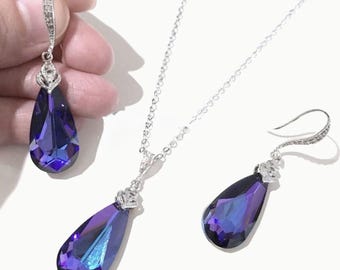 May include: A jewelry set consisting of a silver-toned necklace and two matching drop earrings. Each piece features a large, faceted, pear-shaped deep purple crystal pendant suspended from a silver-toned bail accented with small clear stones.