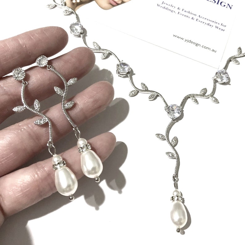 Op de afbeelding: Zilveren ketting- en oorsteensets met een delicaat rankontwerp en glinsterende heldere stenen. Elk stuk is versierd met een parel-druppel.