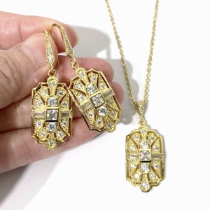 Puede incluir: Un conjunto de joyer&iacute;a en tono dorado que consta de dos pendientes rectangulares colgantes y un collar con colgante a juego. Cada pieza presenta un marco calado intrincado con piedras transparentes, cuadradas y redondas.