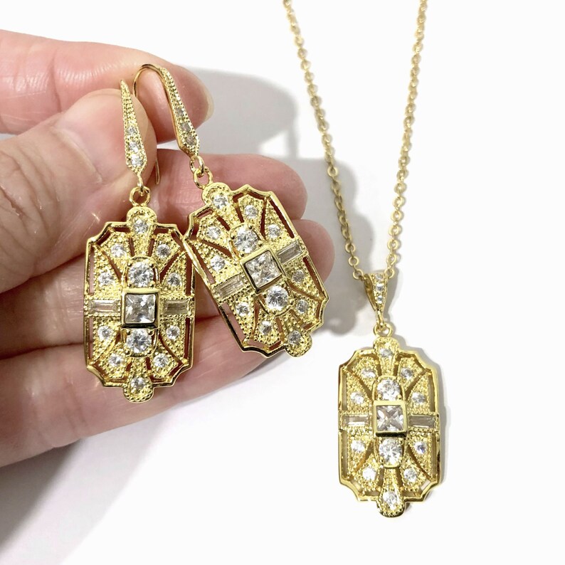 Puede incluir: Un conjunto de joyer&iacute;a en tono dorado que consta de dos pendientes rectangulares colgantes y un collar con colgante a juego. Cada pieza presenta un marco calado intrincado con piedras transparentes, cuadradas y redondas.