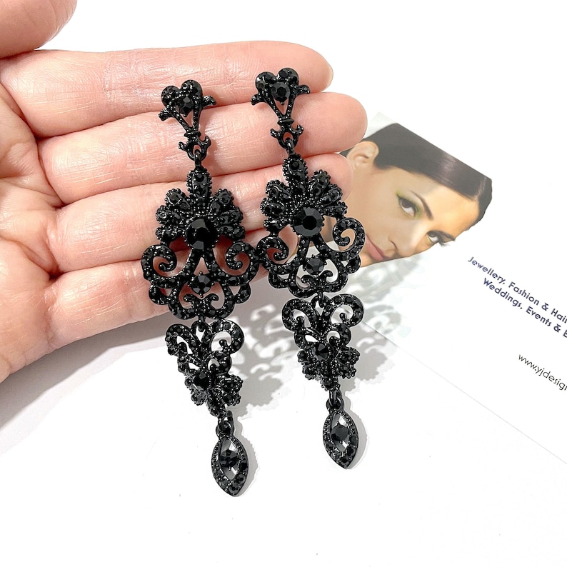 May include: A pair of long, dangling earrings in a deep black finish. Each earring features an intricate, filigree-style metal frame encrusted with small black crystals, a central round black stone, and a teardrop-shaped black crystal at the base.