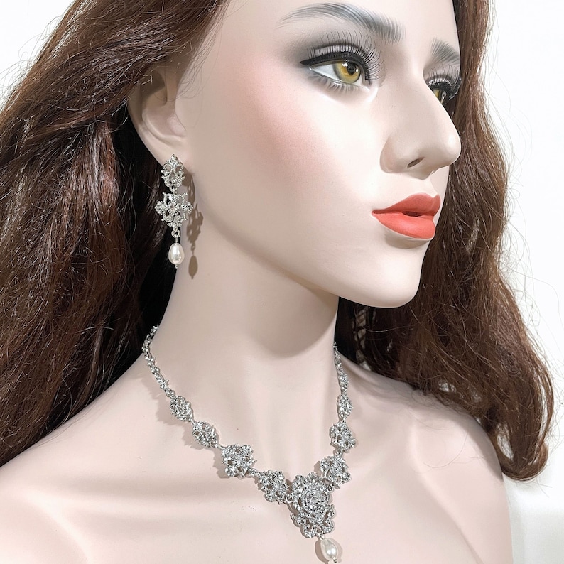 May include: A silver necklace and earrings set with a pearl drop pendant and floral design. The necklace and earrings are adorned with sparkling rhinestones.