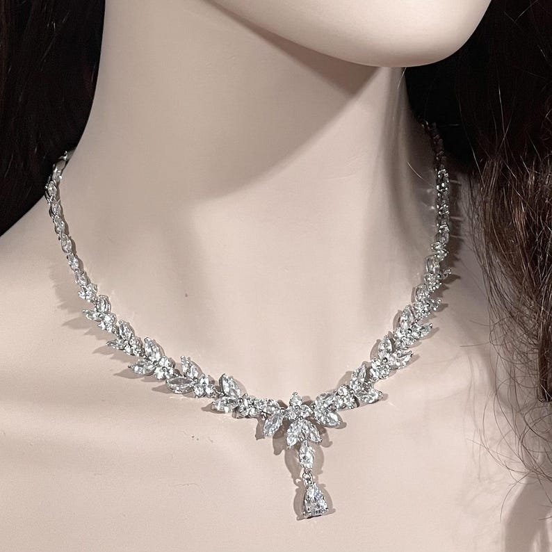 May include: A silver-toned necklace with a leaf-like design, adorned with numerous clear, sparkling crystals. The necklace features a central pendant with a teardrop-shaped crystal. The jewelry is displayed on a mannequin.