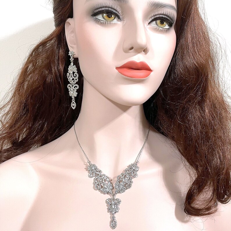 May include: A silver necklace and earrings set with a delicate, intricate design featuring a cascade of sparkling rhinestones.