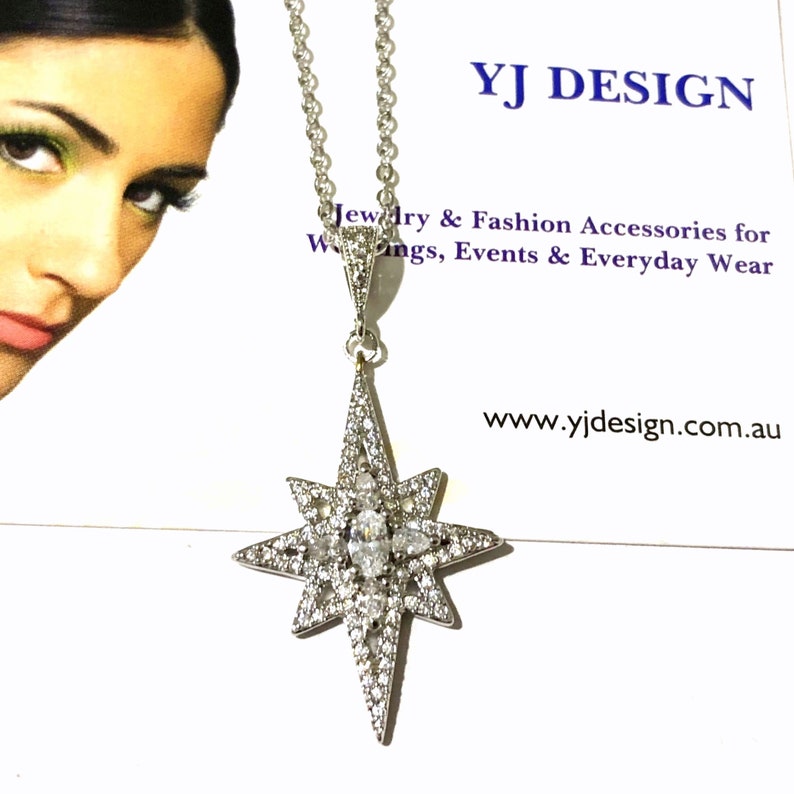 May include: A silver-toned necklace featuring a star-shaped pendant encrusted with numerous small clear stones and a central marquise-cut stone. The pendant hangs from a delicate chain, displayed against a card with the text YJ DESIGN.