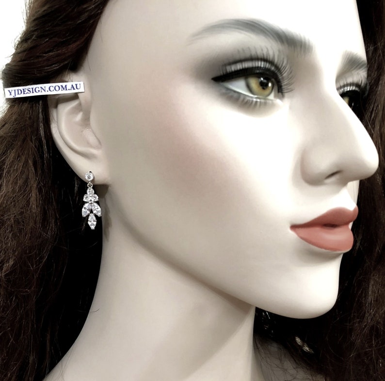 May include: A dangling earring with a cluster of clear, sparkling crystals. The earring features a small, round crystal at the top, with a design of marquise and round-cut crystals cascading downwards. The earring is shown on a mannequin.