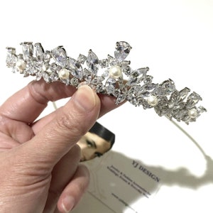 May include: A silver-toned tiara featuring marquise and pear-cut clear crystals arranged in a floral pattern, accented with small round white pearls. The piece is held against a white background with a business card labeled YJ DESIGN.