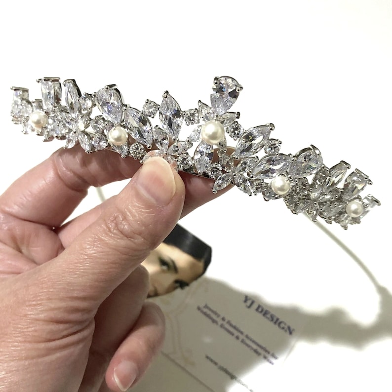 May include: A silver-toned tiara featuring marquise and pear-cut clear crystals arranged in a floral pattern, accented with small round white pearls. The piece is held against a white background with a business card labeled YJ DESIGN.
