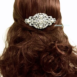 May include: A silver hair comb with a large, ornate design featuring clear rhinestones and white pearls. The comb is shown in a model's brown hair.
