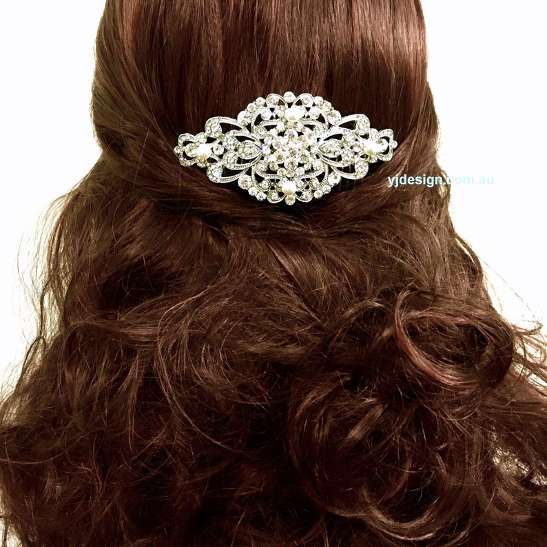May include: A silver hair comb with a large, ornate design featuring clear rhinestones and white pearls. The comb is shown in a model's brown hair.