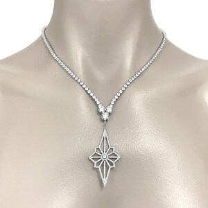 May include: A silver-toned necklace featuring a continuous strand of small, clear, round-cut stones leading to a central cluster of three larger stones. A large, elongated, diamond-shaped pendant with an openwork starburst pattern sits below.