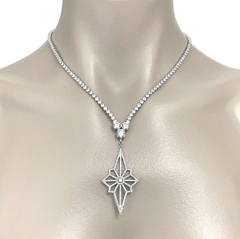 May include: A silver-toned necklace featuring a continuous strand of small, clear, round-cut stones leading to a central cluster of three larger stones. A large, elongated, diamond-shaped pendant with an openwork starburst pattern sits below.