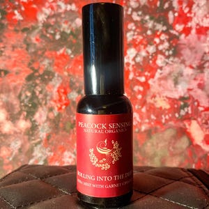 Może przedstawiać: Czarna butelka z czarną nakrętką. Etykieta jest czerwona ze złotym napisem „Peacock Sensing Natural Organics” i „Rolling Into The Deep Aura Mist with Garnet Crystals”. Tło jest czerwone i białe.