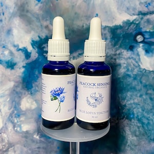 Könnte beinhalten: Zwei dunkelblaue Glasflaschen mit weißen Tropfern. Jede Flasche hat ein weißes Etikett mit blauem Text und einer blauen Lotusblumen-Illustration. Die Etiketten lauten "Peacock Sensing Natural Organics" und "Blue Lotus Tincture 30 ml".