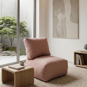 Pode incluir: Uma poltrona moderna de cor rosa empoeirado. A cadeira tem um assento quadrado e uma almofada de encosto grande e de suporte. Uma pequena mesa lateral de madeira com um livro e uma caneca está por perto. A sala tem uma paleta de cores neutras e uma janela grande.