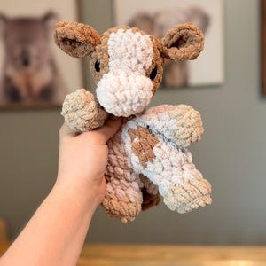 May include: A plush cow toy made of chunky, soft yarn in shades of brown, cream, and white. The stuffed animal features a textured, crocheted appearance with large ears, a rounded snout, and dark, round eyes set into the head.