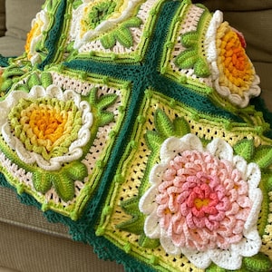 May include: A handmade crochet blanket featuring large, colorful floral designs. The blanket showcases a vibrant palette of pink, yellow, green, and white, with a dark green border. The floral patterns are intricately detailed, creating a cozy and decorative textile.