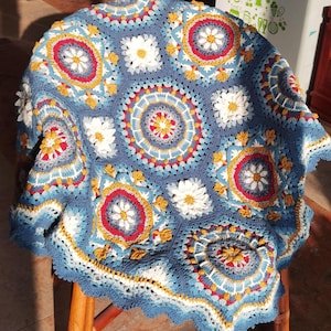 May include: A handmade, blue crochet blanket with a floral pattern. The blanket features white, red, yellow, and gray accents, with a scalloped edge. The blanket is draped over a wooden chair.