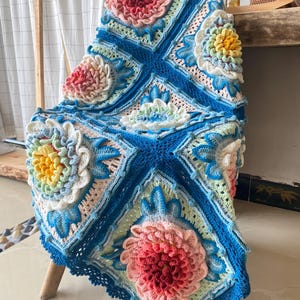 Op de afbeelding: Een handgemaakte, kleurrijke gehaakte deken over een houten stoel. De deken heeft grote, driedimensionale bloemmotieven in roze, geel en blauw, tegen een blauw-witte achtergrond. De randen zijn afgewerkt met een geschulpte rand.