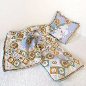 May include: A crocheted blanket and pillow set featuring a daisy and square pattern in shades of white, green, yellow, and brown. The pillow has a light blue background with a daisy and bunny design. The blanket has a light blue and white background.