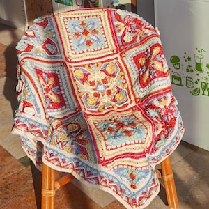 May include: A colorful, handmade crochet blanket draped over a wooden chair. The blanket features a square pattern with red, blue, yellow, and white detailing. The chair has a natural wood finish, and the blanket's texture is clearly visible.