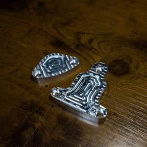 Op de afbeelding: Twee zilverkleurige metalen onderdelen met bewerkte oppervlakken. Het grotere stuk is klokvormig met drie ronde montagegaten, terwijl het kleinere stuk ovaal is met twee montagegaten. Beide zijn voorzien van ingewikkelde, verzonken geometrische patronen.