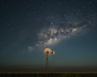 Puede incluir: Un molino de viento de madera con una pala de color marrón y dorado se alza en un campo bajo un cielo nocturno con una Vía Láctea brillante.