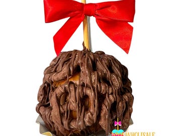 May include: A caramel apple covered in dark chocolate, drizzled with more chocolate, and topped with a bright red bow. The apple sits in a decorative paper cup and is on a wooden stick. The logo for Wholesale Candy Apples is in the bottom right corner.