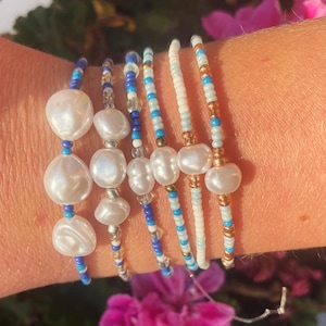 May include: A set of six bracelets featuring white pearls and blue and white beads. The bracelets are layered on a person's wrist.