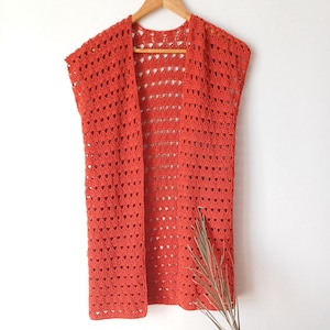 May include: A sleeveless, open-front crochet vest in a warm, reddish-orange hue hangs on a wooden hanger. The open weave design is visible throughout the garment. The vest is displayed against a plain white background, with a sprig of dried foliage in the lower right corner.
