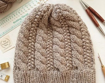 May include: A light brown knit beanie featuring a prominent vertical cable stitch pattern and a ribbed brim. The hat is displayed alongside a knitting gauge tool, a skein of yarn, wooden knitting needles, and a small measuring tape.