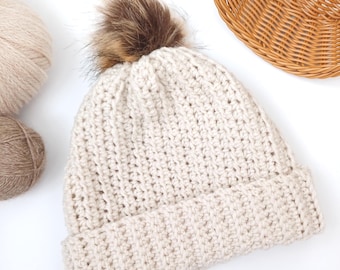 May include: Cream-colored crochet beanie with a ribbed brim and a fluffy brown pom-pom on top. The hat is displayed on a white surface, with two balls of yarn and a woven basket in the background. The beanie is a winter accessory.