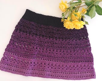 Pode incluir: Uma saia de crochê com um padrão de cores em degradê que transita de um cós canelado preto sólido para um roxo profundo, clareando para um tom violeta vibrante na bainha. A saia é confeccionada com um design de pontos abertos.