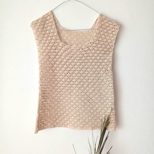 May include: A sleeveless top made of beige yarn featuring an intricate, repeating openwork crochet pattern. The garment has a wide, rounded neckline and is displayed on a metal hanger, showcasing the delicate, textured stitch work throughout.