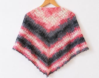 May include: A triangular crochet shawl featuring a chevron pattern with alternating horizontal stripes in shades of cream, bright pink, and dark charcoal gray. The garment is displayed on a wooden hanger, showcasing an open, airy stitch texture.