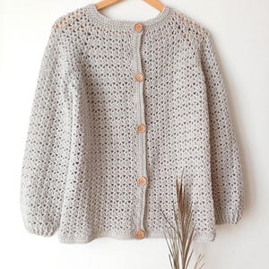 Puede incluir: Un cárdigan de ganchillo de color gris claro con mangas largas y un delicado patrón de puntos calados. La prenda presenta un cuello redondo y una tapeta frontal vertical asegurada con cinco botones redondos de madera clara.