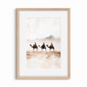 May include: A framed art print showing three figures on camels traversing a desert scene. The artwork uses a muted palette of browns, tans, and whites, with mountains in the background. The print is set within a light-coloured wooden frame.