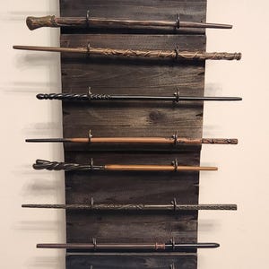 Può includere: Un espositore in legno scuro con diverse bacchette attaccate. Le bacchette, di vari colori e design, sono tenute da ganci di metallo. Le bacchette sembrano essere repliche di una famosa serie fantasy.