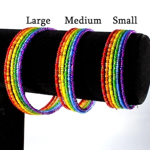 Puede incluir: Tres pulseras de cuentas de arcoíris en diferentes tamaños: grande, mediano y pequeño. Las pulseras están hechas de cuentas de vidrio de colores y se muestran en un soporte negro.