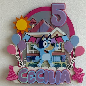 K&ouml;nnte beinhalten: Ein farbenfroher Tortenaufsatz zum Geburtstag mit einer Cartoon-Hundefigur, einem Haus, Luftballons und der Zahl 5. Der Name "CECILIA" wird in gro&szlig;en Buchstaben angezeigt. Der Aufsatz enth&auml;lt eine Sonne, eine Partyh&uuml;tchen und ein Ballon-Tier.