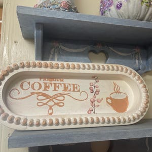 May include: A decorative, off-white wooden tray with the words "PREMIUM COFFEE" in orange script. The tray features an orange coffee cup graphic, a floral design, and a beaded border. The tray is displayed on a blue shelf.