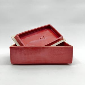 以下が含まれることがあります： 鮮やかな赤色の釉薬で仕上げられた長方形の陶器製ボックス。同色の赤い蓋が斜めに立てかけられています。蓋の内側も光沢のある赤い釉薬で仕上げられており、滑らかな表面が見えています。