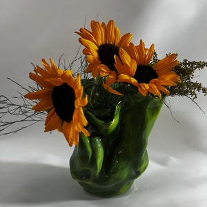 May include: A glossy, organic-shaped green ceramic vase with a fluid, rippled surface texture. The vase holds a bouquet of three bright yellow sunflowers with dark brown centers, accompanied by thin, wispy green stems and small dried foliage.