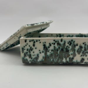 Może przedstawiać: Prostokątne ceramiczne pudełko z pokrywką, o białym kolorze bazowym i nakrapianym wzorze z ciemnoszarej i turkusowej glazury. Pokrywka jest częściowo otwarta, ukazując wewnętrzne ścianki pudełka o tym samym nakrapianym wykończeniu.