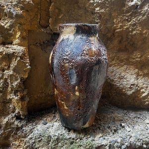 May include: A ceramic vase with a heavily textured, uneven surface featuring a mottled color palette of dark brown, charcoal black, and hints of cream. The vessel has a wide, rounded body that tapers toward a narrow, open circular rim at the top.