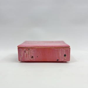 May include: A rectangular ceramic box with a lid, featuring a pink glaze with subtle variations in tone and small dark speckles throughout the surface. The box has a smooth, flat top and straight sides, showcasing a handcrafted finish.