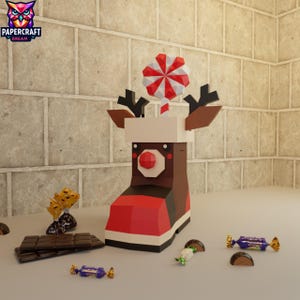 Puede incluir: Una bota de reno de papel con un bastón de caramelo rojo y blanco y una nariz roja. Barras de chocolate y caramelos envueltos rodean la bota, creando una escena festiva navideña.