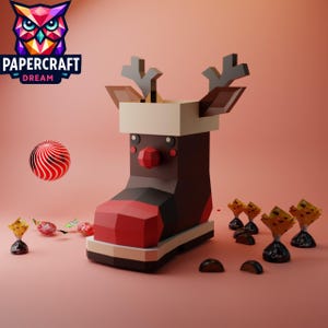 Könnte beinhalten: Ein roter und brauner Papiercraft-Rentier-Stiefel mit weißem Schaft und schwarzen Geweih. Der Stiefel steht auf einer rosa Oberfläche mit einem rot-weiß gestreiften Ball und mehreren verpackten Pralinen.